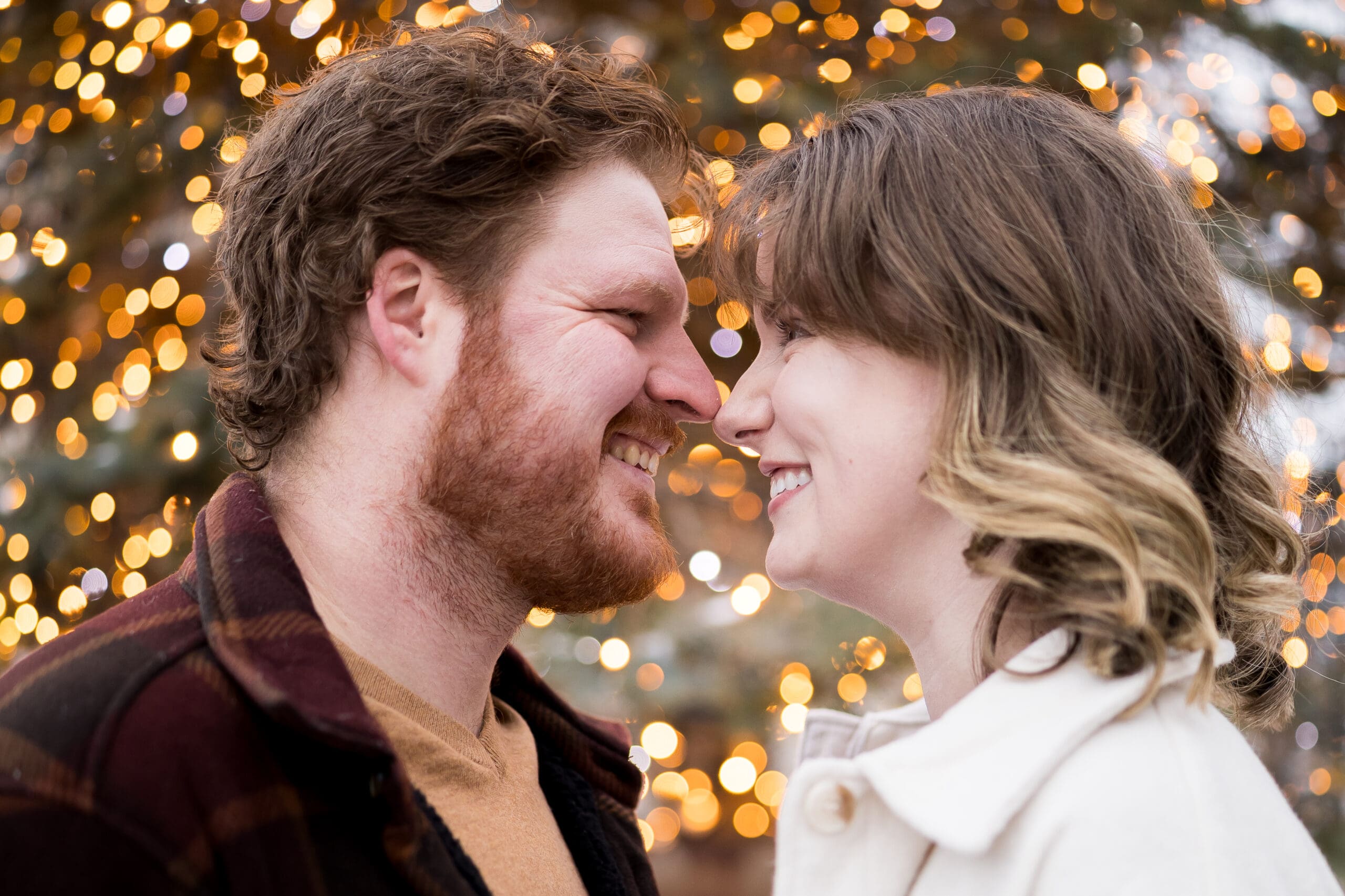 an engaged couple nuzzles their noses in front of a tree with lights